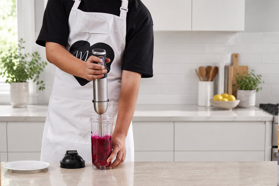 A person wearing a white apron over a black shirt uses an immersion blender to mix a dark magenta-purple liquid in a clear blending cup on a kitchen counter. The immersion blender has a silver and black handle, and the cup is filled with the dark purple mixture.