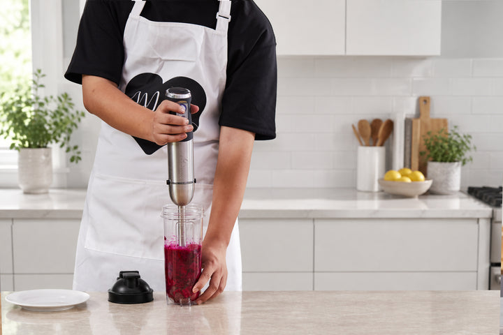 A person wearing a white apron over a black shirt uses an immersion blender to mix a dark magenta-purple liquid in a clear blending cup on a kitchen counter. The immersion blender has a silver and black handle, and the cup is filled with the dark purple mixture.